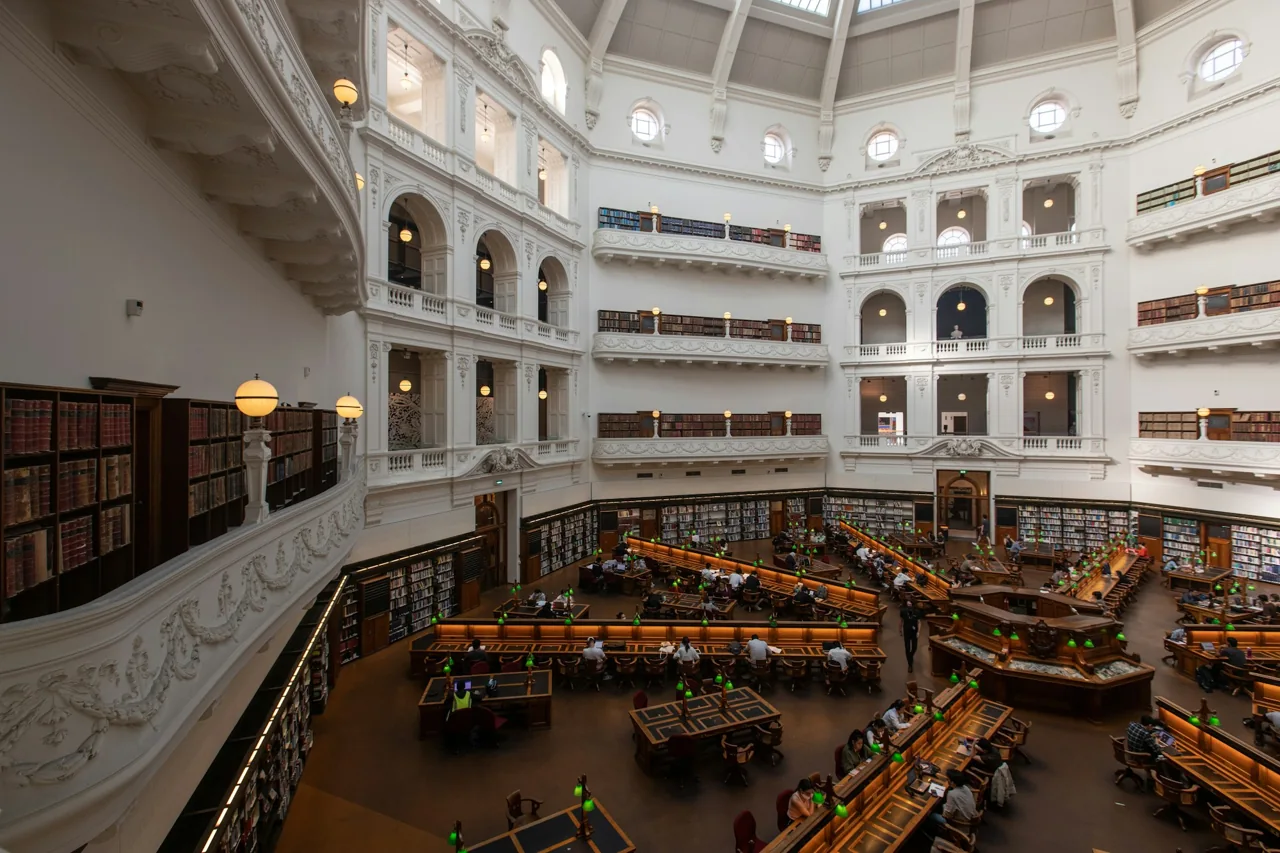 A spacious library reading room with tiers of bookshelves and study desks.